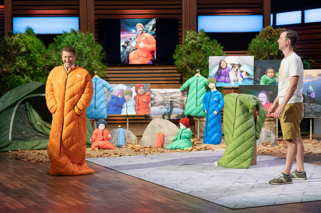 Two people wearing colorful sleeping bags on a stage with a backdrop of people in similar sleeping bags.
